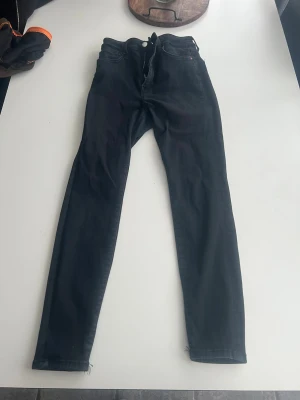Svarta Peachy High Waist jeans - Säljer ett par svarta Peachy High Waist jeans i storlek S. De har hög midja och ankellångt snitt med smal passform. Klassisk femficksmodell och dragkedja med knapp framtill. Perfekta till sneakers eller boots.
