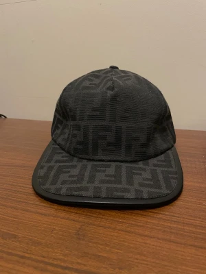 Svart keps från Fendi med FF-mönster - Säljer en svart keps från Fendi med det klassiska FF-monogrammet över hela kepsen. Kepsen har böjd skärm med svart kant och är tillverkad i tyg med exklusiv känsla. Perfekt för dig som gillar stilrena accessoarer med lyxig touch.