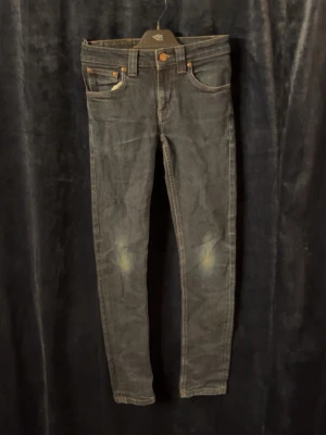 Nudie Jeans - Snygga blåa jeans från Nudie Jeans med klassiska fem fickor och guldfärgade sömmar. Jeansen har en något tvättad look och detaljerade bakfickor med Nudie-loggan. Perfekta för en trendig och avslappnad stil. Skriv för frågor (storlek 24/32) (passar folk som är ca 145-160 cm lång