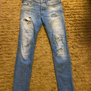Slim fit blå slitna jeans från Jack & Jones - Säljer ett par ljusblå slim fit jeans från Jack & Jones med slitningar och hål på benen för en trendig look. Jeansen har klassisk femficksmodell, knappgylf och är tillverkade i bomullsmix med stretch. Perfekta för dig som gillar en avslappnad men snygg stil.