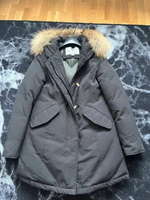 Mörkgrå parkas med pälskrage från Marville - Snygg mörkgrå parkas från Marville med fluffig pälskant på huvan och quiltat foder. Jackan har dragkedja, knappar och stora fickor framtill. Perfekt för kalla dagar och ger en riktigt stilren look.