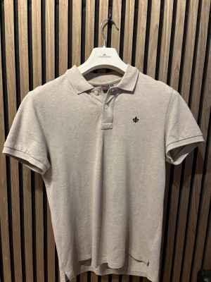 Beige pikétröja från Morris - Klassisk beige pikétröja från Morris med korta ärmar och krage. Tröjan har knappar framtill och en liten broderad logga på bröstet. Tillverkad i mjuk bomull som känns skön mot huden. Perfekt till jeans eller shorts.