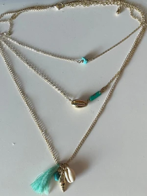 Guldigt halsband med snäckor och tofs - Säljer ett trendigt lager-på-lager halsband i guldton med turkosa pärlor, snäckor och en liten tofs. Perfekt accessoar för dig som gillar boho-stil och strandvibbar. Halsbandet har flera hängen, bland annat snäcka, tofs och små pärlor.