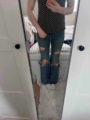 Blå bootcut jeans med slitningar - Säljer ett par blå bootcut jeans med stora slitningar över knäna och råa kanter. Jeansen har en klassisk femficksdesign och är tillverkade i denim. Perfekta för dig som gillar en avslappnad och trendig look.