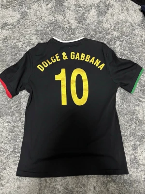 Svart Dolce & Gabbana t-shirt med nummer 10 - Svart t-shirt från Dolce & Gabbana med guldfärgat nummer 10 på både fram- och baksida samt logga på bröstet. Kontrasterande vita, gröna och röda detaljer vid hals och ärmslut. Tillverkad i 100% bomull, perfekt för dig som gillar sportig stil och snygga detaljer.