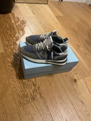 Philippe Model blå/grå sneakers - Säljer ett par Philippe Model sneakers i blått och grått med beige detaljer. Skorna har snörning, rund tå och platt sula. Ovandelen är i mocka och textil, med vita och grå inslag samt en snygg kontrast på sulan. Perfekta för dig som gillar stilrena och sportiga sneakers.