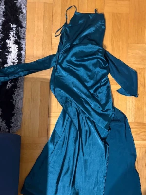 Lång blå satinklänning med öppen rygg - Snygg långklänning i glansigt blått satinmaterial med smala axelband och öppen rygg. Klänningen har knyt i ryggen och en elegant, svepande siluett som ger en lyxig känsla. Perfekt för dig som vill sticka ut med en stilren och trendig look.