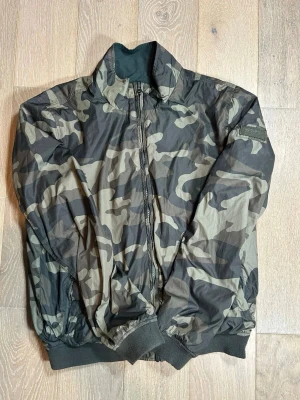 Camouflage Woolrich jacka  med dragkedja  - Snygg bomberjacka i camouflage-mönster med hel dragkedja framtill och ribbade muddar vid ärmslut och midja. Jackan har klassisk bomberkrage och är perfekt för dig som vill sticka ut med en streetstyle-look.