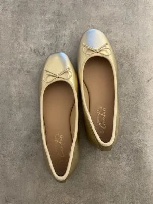 Guldiga ballerinaskor  - Snygga ballerinaskor i guld från New Look med rund tå och dekorativ rosett framtill. Skorna är aldrig använda.