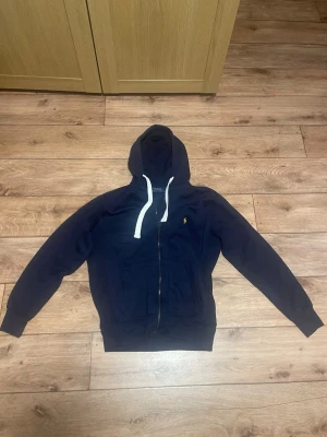 Mörkblå hoodie från Polo Ralph Lauren - Snygg mörkblå hoodie med dragkedja från Polo Ralph Lauren. Klassisk design med vit dragsko i huvan och den ikoniska lilla gula loggan på bröstet. Perfekt för en avslappnad och stilren look. Mjuk bomullskvalitet och två fickor framtill.