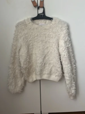 Vit fluffig stickad tröja - Supermjuk och fluffig vit stickad tröja med rund hals och långa ärmar. Tröjan har en oversized känsla och ribbade muddar vid ärmslut och nederkant. Perfekt för dig som gillar mysiga och trendiga plagg.