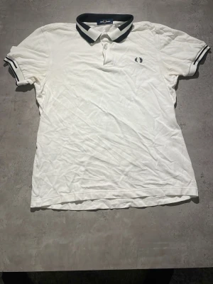 Vit pikétröja från Fred Perry - Klassisk vit pikétröja från Fred Perry med svart krage och svarta detaljer på ärmsluten. Tröjan har knappslå framtill och den ikoniska Fred Perry-loggan broderad på bröstet. Tillverkad i bomull för en skön och luftig känsla.