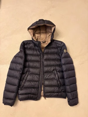 Svart dunjacka med huva från Colmar (dam) - Snygg svart dunjacka från Colmar med huva och quiltad design. Jackan har dragkedja framtill, två sidofickor med dragkedja och en logotyp på ärmen. Perfekt för kalla dagar. Materialet är polyester och dun. Jackan är i storlek 40.