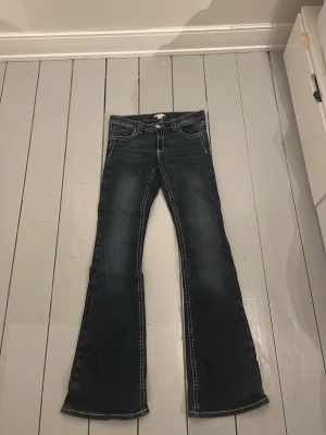 Blå bootcut jeans med broderade fickor - Säljer ett par blå bootcut jeans med snygga broderade detaljer på bakfickorna. Jeansen har kontrastsömmar och en fyrficks design. Materialet är jeans i bomull och passformen är normal med utsvängda ben.