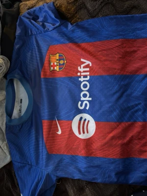FC Barcelona Nike matchtröja blå/röd - Säljer en officiell FC Barcelona matchtröja från Nike med Dri-FIT material. Tröjan är blå och röd med klubbmärke, Nike-logga och Spotify som sponsor framtill. Perfekt för dig som älskar fotboll och vill bära ditt favoritlag på match eller träning.