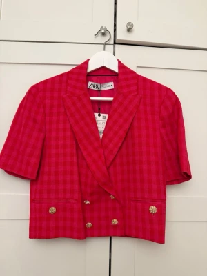 Rutig rosa kavaj från Zara - Kort rutig kavaj i stark rosa från Zara med dubbelknäppning och guldfärgade knappar. Kavajen har korta ärmar, breda slag och en croppad passform. Perfekt för dig som vill sticka ut med en färgstark look. Prislapp kvar, aldrig använd! 