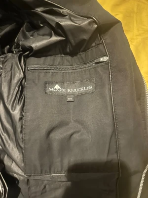 Svart dunjacka Moose Knuckles - Snygg svart dunjacka från Moose Knuckles med stor huva och vit fuskpäls. Jackan har dragkedjor i sidorna, quiltad design och logga på ärmen. Perfekt för kalla dagar och riktigt varm. Klassisk pufferstil med lång ärm.