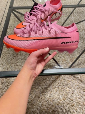 Nike Air Zoom rosa fotbollsskor - Säljer ett par Nike Air Zoom fotbollsskor i en snygg rosa färg med orange detaljer och svart swoosh. Skorna har snörning och är tillverkade i syntetmaterial för bästa prestanda på planen. Perfekta för dig som vill sticka ut på fotbollsplanen!