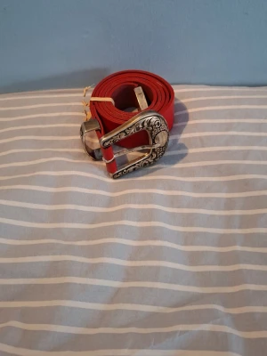 Rött bälte med dekorerat spänne - Säljer ett rött bälte med ett snyggt, silverfärgat spänne med detaljerade mönster. Perfekt accessoar för att lyfta din outfit och ge en cool touch. Bältet är i skinn och har en klassisk form.