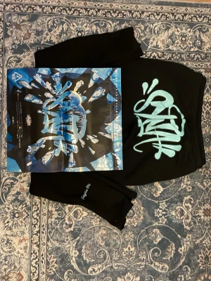 Svarta mjukisbyxor med blått tryck - Säljer ett par svarta mjukisbyxor med stort blått graffiti-inspirerat tryck på baksidan. Byxorna har en relaxed passform och är perfekta för chill dagar. Materialet känns mjukt och skönt, troligen bomull eller polyester. Grym streetwear-vibe!