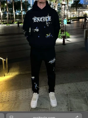 Svarta mjukisbyxor med tryck och färgstänk - Svarta mjukisbyxor från Excite med coolt tryck på ena låret och färgstänk i blått, gult och vitt. Byxorna har snörning i midjan och en slim passform. Perfekta för chill dagar och streetwear-stil.