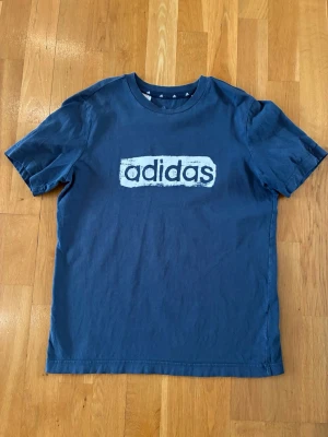 Blå Adidas t-shirt med logga - Snygg blå t-shirt från Adidas med stor vit logga framtill. Klassisk rund halsringning och korta ärmar. Tillverkad i mjuk bomull som känns skön mot huden. Perfekt för en avslappnad och sportig stil.