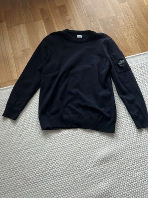 Svart stickad tröja från C.P. Company - Snygg svart stickad tröja från C.P. Company med rund halsringning och ribbade muddarstorlek 52 med andra ord L. Tröjan har en ikonisk detalj med logga på ärmen och är tillverkad i mjukt material som passar perfekt till en avslappnad stil. Mycket hög kvalité och använd max 5 gånger. DU VET VARFÖR DEN ÄR BILLIG!