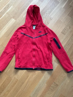 Röd hoodie från Nike med dragkedja - Snygg röd hoodie från Nike med hel dragkedja framtill och svarta detaljer. Hoodien har en huva, svarta linjer över bröstet och en praktisk ficka med dragkedja på ena ärmen. Perfekt för en sportig och avslappnad stil.
