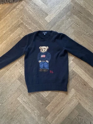 Mörkblå stickad tröja Polo Bear RL - Säljer en mörkblå stickad tröja från Ralph Lauren med den ikoniska Polo Bear på bröstet. Tröjan har rund halsringning, långa ärmar och ribbade muddar. Björnen har en amerikansk flagga på tröjan och RL-broderi i rött nedtill. Perfekt för dig som gillar klassisk preppy stil.