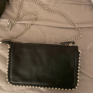 Svart kuvertväska med kedja och nitar - Stilren svart kuvertväska i skinn med silverfärgad kedja och coola nitar längs kanterna. Perfekt accessoar för att lyfta din outfit med en edgy touch. Väskan har dragkedja upptill och plats för det viktigaste.