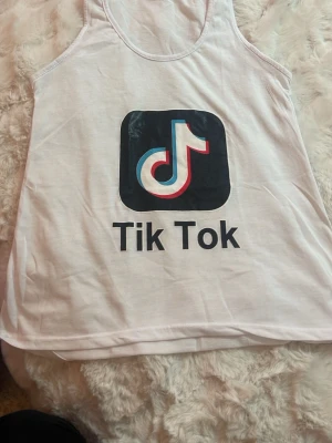 Vit TikTok linnetopp - Vit linneteöja med stort TikTok-tryck framtill. Toppen är ärmlös med rund halsringning och har en avslappnad passform. Perfekt för dig som vill visa din kärlek till TikTok och ha en enkel, trendig stil. 