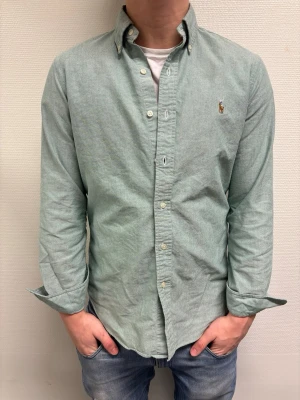 Grön skjorta från Polo Ralph Lauren - Klassisk grön skjorta från Polo Ralph Lauren med button-down krage och broderad logga på bröstet. Skjortan har långa ärmar och knappar framtill. Perfekt till jeans eller chinos för en clean look.