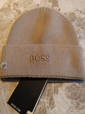 Beige ribbstickad mössa från BOSS - Den är helt ny oanvänd med etikett på. Snygg beige ribbstickad mössa från BOSS med uppvikt kant och broderad logga framtill. Klassisk och stilren design som passar perfekt till höstens och vinterns outfits. Tillverkad i mjukt material för extra komfort.