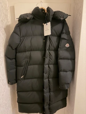  Moncler jacket - Snygg svart lång dunjacka från Moncler med hög krage och huva. Jackan har quiltad design, dragkedja och knappar framtill samt Moncler-logga på ärmen. Perfekt för kalla vinterdagar och riktigt varm. Klassisk pufferstil med praktiska fickor. Helt ny led taggar kvar