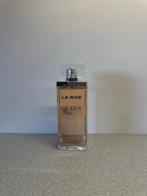 La Rive Queen of Life parfym - La Rive Queen of Life Eau de Parfum i en elegant, rektangulär glasflaska med transparent lock och ljusrosa doftvätska. Flaskan har stilren design med svart text och silverdetaljer. Perfekt för dig som gillar feminina och fräscha dofter. ”sensuella blomdofter och varma tränoter”💐
