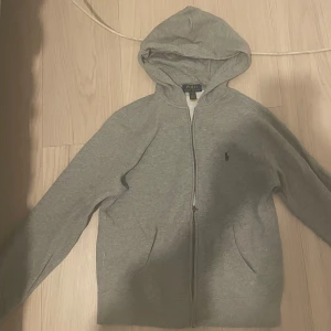 Grå zip hoodie från Polo Ralph Lauren - Snygg grå hoodie med dragkedja från Polo Ralph Lauren. Klassisk design med huva, två fickor framtill och den ikoniska lilla loggan broderad på bröstet. Perfekt till chill vardagsstil och enkel att matcha med allt. Mjuk bomullskänsla.