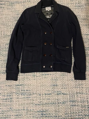 Mörkblå kavaj från Woolrich - Snygg mörkblå kavaj från Woolrich med dubbelknäppning och två framfickor. Klassisk krage och långa ärmar. Tillverkad i mjuk bomull som ger en avslappnad men stilren look. Storlek L men passar XS