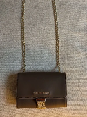 Brun/taupe axelrämsväska från Mario Valentino - Snygg brun/taupe axelrämsväska från Mario Valentino med guldfärgad kedjerem och dekorativ tofs i metall framtill. Väskan har ett stilrent och elegant utseende med metallogo på framsidan. Perfekt storlek för det viktigaste och tillverkad i slätt skinnliknande material.