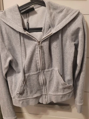 Grå zip hoodie från Juicy Couture - Säljer en klassisk grå zip hoodie från Juicy Couture med broderad JC-logga på bröstet och dragkedja med snygg JC-detalj. Hoodien har huva med dragsko och två fickor framtill.