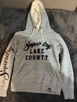 Grå Superdry hoodie med text - Snygg grå hoodie från Superdry med stor broderad text 'Superdry Lake County' på bröstet och 'Superdry' längs ena ärmen. Mjuk fleecefodrad huva med vita dragsnören och känguruficka framtill. Perfekt för chill dagar och streetwear vibes. Tröjan är storlek xs