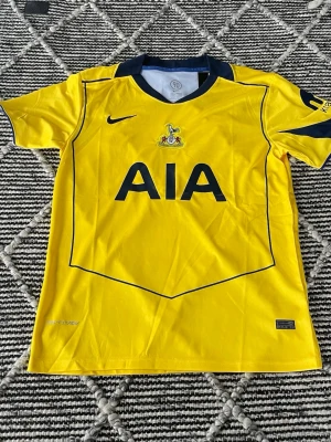 Gul Tottenham Hotspur matchtröja Nike - Snygg gul Tottenham Hotspur matchtröja från Nike med mörkblå detaljer och klubbmärke på bröstet. Tröjan har AIA-logga framtill och korta ärmar. Tillverkad i lätt och ventilerande material, perfekt för fotboll eller träning. Ny med prislapp
