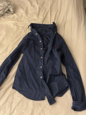 Blå slim fit skjorta från Ralph Lauren - Säljer en mörkblå skjorta från Ralph Lauren i slim fit med klassisk krage och knappar framtill. Skjortan har det ikoniska röda Polo-broderiet på bröstet och är gjord i tunn bomullstwill. Perfekt till jeans eller chinos för en clean look.