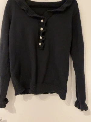 Svart stickad tröja med pärlknappar - Supersöt svart stickad tröja med volangkant vid halsen och ärmsluten. Framsidan har dekorativa pärlknappar och en snygg ribbad struktur. Vet inte vart den är från då lappen inte sitter kvar.