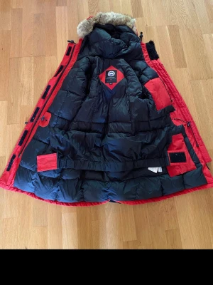 Röd dunjacka från Canada Goose XS - Säljer en röd och svart dunjacka från Canada Goose i storlek XS. Jackan har huva med päls, flera fickor och detaljer i svart. Materialet är polyester och dun, perfekt för kalla dagar. Klassisk Canada Goose-logga på ärmen och insidan.