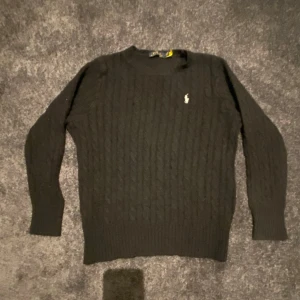 Svart stickad tröja från Polo Ralph Lauren - Svart stickad tröja från Polo Ralph Lauren med klassiskt kabelstickat mönster och broderad logga på bröstet. Tröjan har rund halsringning och långa ärmar.