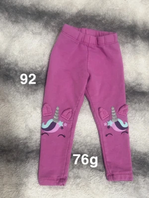 Rosa leggings med enhörningsmotiv - Säljer ett par söta rosa leggings med enhörningsmotiv på knäna. Byxorna har glittriga horn, 3D-öron och stjärndetaljer i lila, turkos och silver. Perfekta för dig som gillar färgglada och lekfulla plagg.🌸 Fint använt skick. Storlek 92