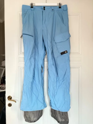 Ljusblå snowboard/skidbyxor, BURTON, Herr stor. L - Säljer ett par herr ljusblå täckbyxor från BURTON i storlek Large. Byxorna har flera fickor med dragkedja och lock, justerbar midja och snölås vid bensluten. Materialet är slitstarkt. Säljer pga vinter garderobsrensning. Fint färg men ändå lätt att se på backen. 
