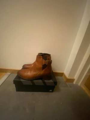 Bruna Daisa boots i skinn - Säljer Snygga bruna Daisa boots i skinn med klassisk rund tå och låg klack. Bootsens design har en cool spänndetalj på sidan och dragkedja på insidan för enkel påtagning. Perfekta för dig som vill ha en stilren och tidlös look.