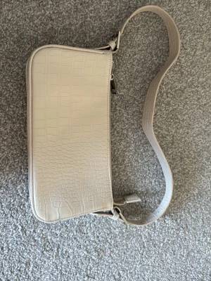Beige axelväska i croco-imitation - Säljer en beige axelväska i syntetmaterial med croco-mönster. Väskan har en stilren, rektangulär form och stängs med dragkedja upptill. Perfekt storlek för det viktigaste och enkel att matcha till olika outfits.