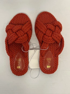Roströda sandaler i rep från H&M - Säljer ett par unika sandaler från H&M i storlek 35. De är gjorda i ett tjockt, flätat repmaterial i en snygg roströd färg. Sandalerna har öppen tå och platt sula, perfekta för sommarens alla dagar.